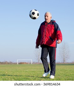 Old Man In Seventies Kicking A Soccer Ball On The Grass Field