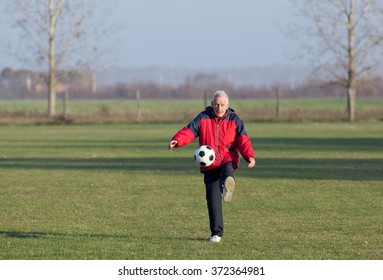 Old Man In Seventies Kicking A Soccer Ball On The Grass Field