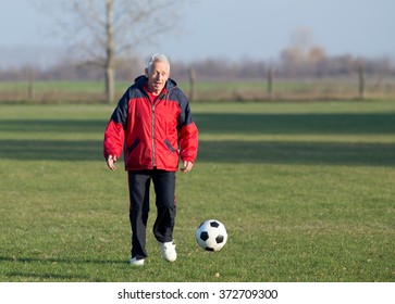 Old Man In Seventies Kicking A Soccer Ball On The Grass Field