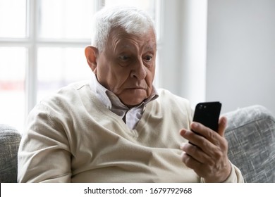 Old man sit on sofa hold smartphone look at device screen feels confused shocked by received sms message. Older generation and modern gadgets apps usage difficulties, broken cell, need repair concept - Shutterstock ID 1679792968