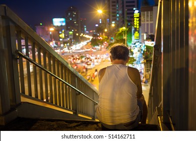 Old man sits on walking bridge seeing crowded city by night. Concept of lonely old man 