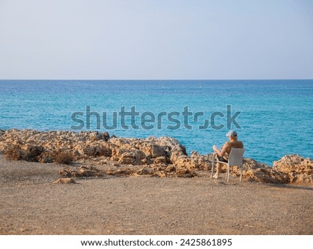 Old man sitting in a chair with a notebook and a pen, having rest and sunbathing at the Nissi beach with beautiful porous rocks near blue Mediterranean sea in Ayia Napa, Cyprus.