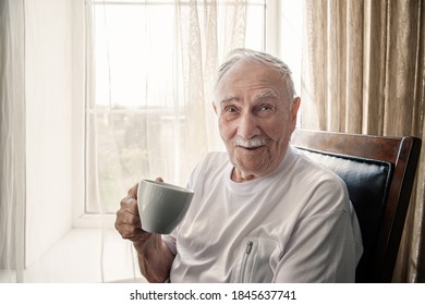 Old Man Is Sitting In A Chair. A Smiling Old Man Is Sitting In A Chair By The Window With A Cup Of Coffee Or Tea. Portrait, Close-up, Copy Space