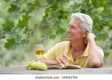 Old Man Sitting At A Table Drinking Wine And Reading A Book