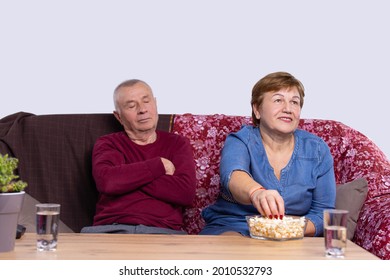 An Old Man Sleeping On A Couch, His Old Wife Eating Popcorn And Watching Tv-set With Two Glasses Of Water At A Table.