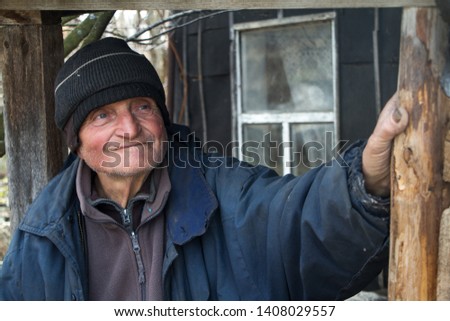 An old man in sloppy clothes stands on the threshold of his own ruined house and looks into the distance, loneliness in the village, life beyond poverty