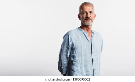 Old Man Standing With His Hands Behind Him Looking At Camera. Portrait Of A Senior Man Standing Against White Background.