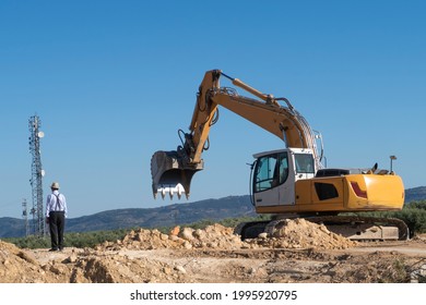 An Old Man Staring At A Work On A Ground Of Disturbed Earth: An Excavator With Chains Preparing The Bucket To Catch Earth. In The Background A Cell Tower And A Mountain With Vegetation