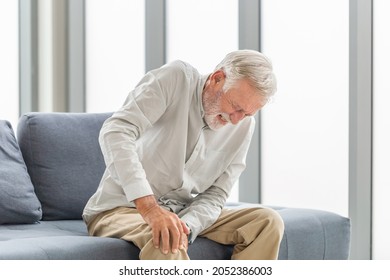 Old man suffering from knee pain sitting sofa in the living room, Elderly man suffering from knee pain while sitting on the sofa