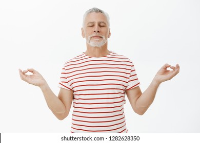 Old Man Taking Deep Breath Peacefully Meditating Over Gray Background. Portrait Of Determined And Calm Good-looking Senior Male With Grey Beard Standing In Lotus Pose With Closed Eyes Practicing Yoga