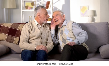 Old man talking to deaf friend sitting on sofa at home, hearing disease, problem