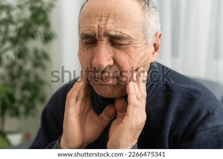 Old man with toothache. Elderly senior man has toothache. Unhappy man face in tooth pain sitting on sofa at home, feel sick unwell. Sad aged man hand holding his chin. Adult suffering toothache