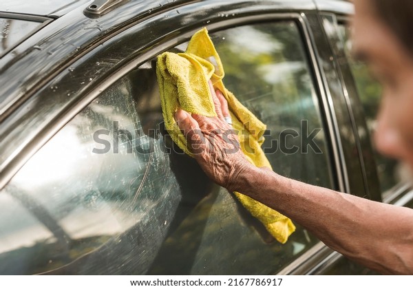 An old man uses a yellow washcloth to clean the\
window of his black hatchback. A car owner cleaning and taking care\
of his auto in the morning.
