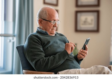 Old Man Wearing Eyeglasses To Use Smartphone At Home. Handsome Senior Man At Home Relaxing On Couch Reading A Message On Cellphone. Senior Sitting Alone In His Living Room And Using Mobile Phone.