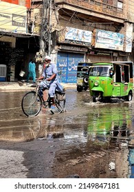 old man with white beard riding a bicycle on a flooded street. Tuk Tuk in green color. Multan, Pakistan. 04. 12. 2022.