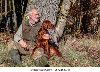 Old man who loves nature, sits in the beautiful spring sun outside in the forest at a tree and holds his young Irish Setter in his arm, who watches the area carefully.