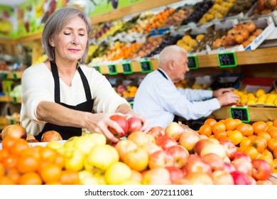 Old Man And Woman In Aprons Working In Salesroom Of Greengrocer. They're Setting Out Fruits On Shelves..