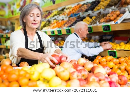 Old man and woman in aprons working in salesroom of greengrocer. They're setting out fruits on shelves..