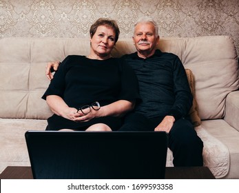 Old Man And Woman In Black Clothes Are Sitting At Home In Front Of A Laptop.