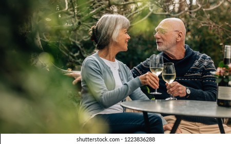 Old Man And Woman Spending Time Together Drinking Wine. Old Couple Sitting Together In A Garden Holding Wine Glasses Looking At Each Other.