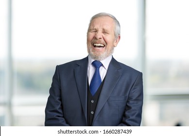 Old manager in office laughing hard. Cheerful boss in business suit portrait. Blurred windows background.