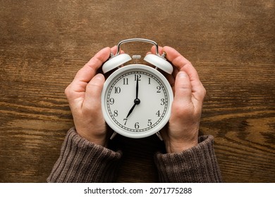 Old mature woman hands holding white alarm clock on dark brown wooden table background. Closeup. Point of view shot. Seven o'clock. Concept of female biological time. - Powered by Shutterstock