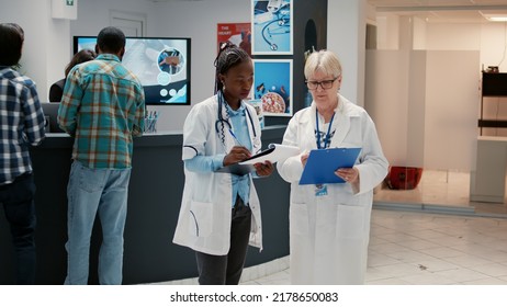 Old Medic And African American Physician Analyzing Checkup Papers In Waiting Room Lobby At Hospital Reception. Diverse Medical Staff Having Healthcare Appointments With Patients.