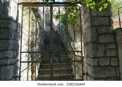 Old metal entrance gateway or gate with door lock leading to steep stairs. Gateposts are consisting of white stones equally walled. Entrance is in shadow and overgrown with green plants and leaves.