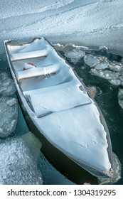 An Old Metal Fishing Boat Moored At A Fishing Wharf.  The Open Boat Is Covered In Fresh White Snow. The Small Row Boat Is Sinking From The Weight Of Snow And Ice. The Ocean Surrounding Is Frozen Ice  