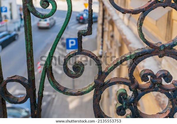 Old metal lattice of a balcony with traces of rust\
and old paint over the street of the old city, along which cars\
drive on a sunny day