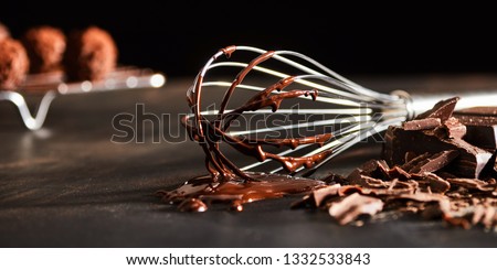 Old metal whisk coated in melted chocolate lying on a kitchen counter alongside chopped chocolate candy in banner format with copy space