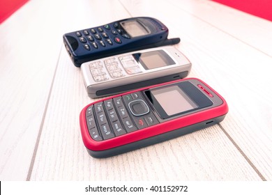 Old Mobile Phone Of Different Colors Lined Up In Vertical Perspective - Cell Devices Retro Technology Disposed On Wooden Panel - Close Up Cellphones Outdated In Showcase Side View - Old Tech Display