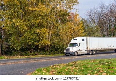 Old model of white American bonnet used big rig semi truck with shabby dry van semi trailer driving on straight sunny autumn road with yellow and green trees and fallen leaves - Shutterstock ID 1297874314