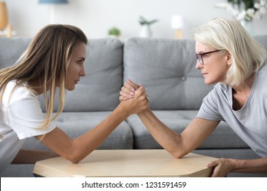 Old mother and grown millennial daughter in law arm wrestling having fight or family conflict, young and senior women rivals competing, disagreement and different generations problems concept
