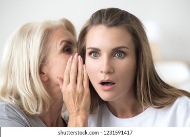 Old mother whispering in ear of adult daughter telling secret or unbelievable news, older and young women gossiping, shocked lady surprised by hearing latest rumors chatting with female aged friend