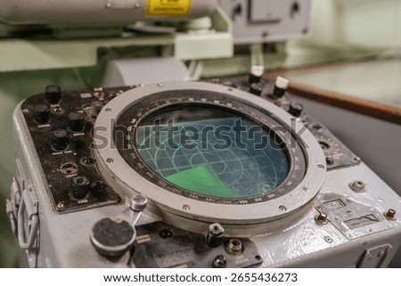 Old naval radar console with a circular green screen and mechanical controls. The radar shows a scanning grid used for detecting objects at sea.