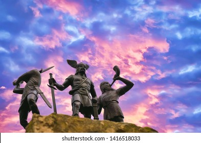 Old Norse God Odin standing on a hard rock with two Vikings, one of them is trumpeting with horn and the other raised a sword (toy plastic soldiers), burning cloudy sky, Valhalla and Ragnarok themes 