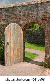 Old open arched wooden door set into an old red brick wall and leading to a grassed area beyond.