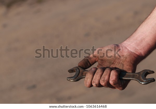 An old open-ended wrench in the hand. Working\
hands in mud, soot, dust, oil. An experienced car service master.\
Can be used as a background for an inscription, cover or part of a\
design.