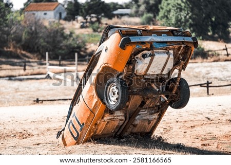 Old orange car flipped vertically, partially buried in dry dirt, in a rural setting with a blurred house and wooden fence in the background