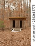 Old outside in the woods an in-ground spring fed white baptistry. Small building with two doors behind.  Isolated nestled shade with trees and brown dry leaves on the ground on a cloudy day. No people