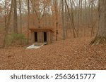 Old outside in the woods an in-ground spring fed white baptistry. Small building with two doors behind.  Isolated nestled shade with trees and brown dry leaves on the ground on a cloudy day. No people