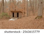 Old outside in the woods an in-ground spring fed white baptistry. Small building with two doors behind.  Isolated nestled shade with trees and brown dry leaves on the ground on a cloudy day. No people