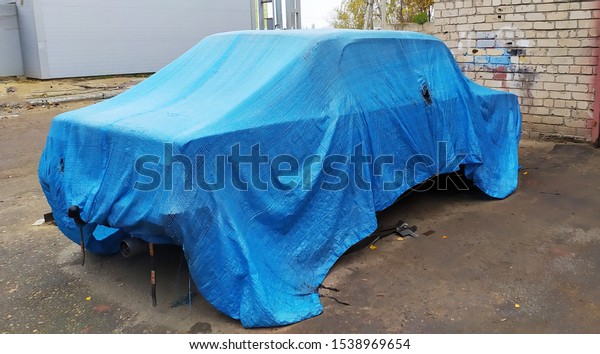 Old Passenger car covered with a blue protective\
sheet in street outdoor