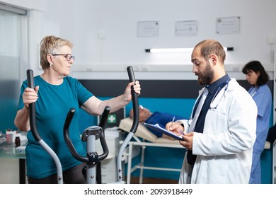 Old Patient Doing Physical Activity With Stationary Bicycle While Doctor Giving Assistance For Physiotherapy. Retired Woman Using Bike For Fitness Workout To Recover From Injury.