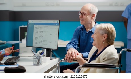 Old Patient With Physical Disability Doing Checkup Visit With Medic And Husband, Talking About Disease Diagnosis. Doctor Consulting Woman Wheelchair User At Healthcare Exam Appointment.