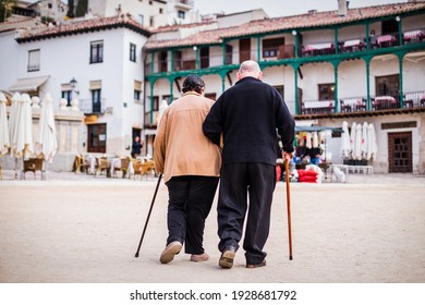 Old Pensioner Couple Walking Together. Elderly Man And Woman Travel To Chinchon In Spain With Love. A Traditional Travel Famous Travel Destination With Typical Spanish Balcony Facades.