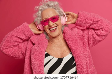 Old people, entertainment and fun concept. Positive good looking woman pensioner catches rhythm of music, wears headphones, enjoys favorite song, dressed in fashionable clothes, trendy sunglasses