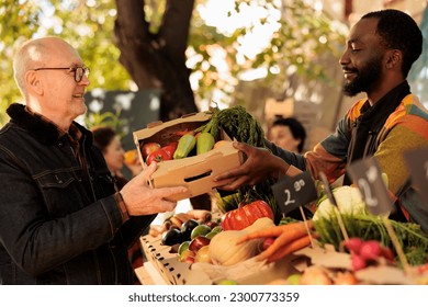 Old person receiving box of fresh fruits and veggies at farmers market, visiting stand on sunny day. Smiling african american man small business owner selling home grown organic produce. - Powered by Shutterstock