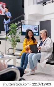 Old Physician Consulting Asian Patient At Health Center, Analyzing Disease Report Using Digital Tablet. Senior Doctor Talking To Person With Disease Diagnosis At Consultation In Hospital Waiting Room